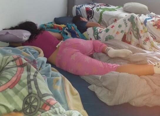 A person is sleeping in a bed, covered with blankets. Another person is partially visible in the background, also sleeping.