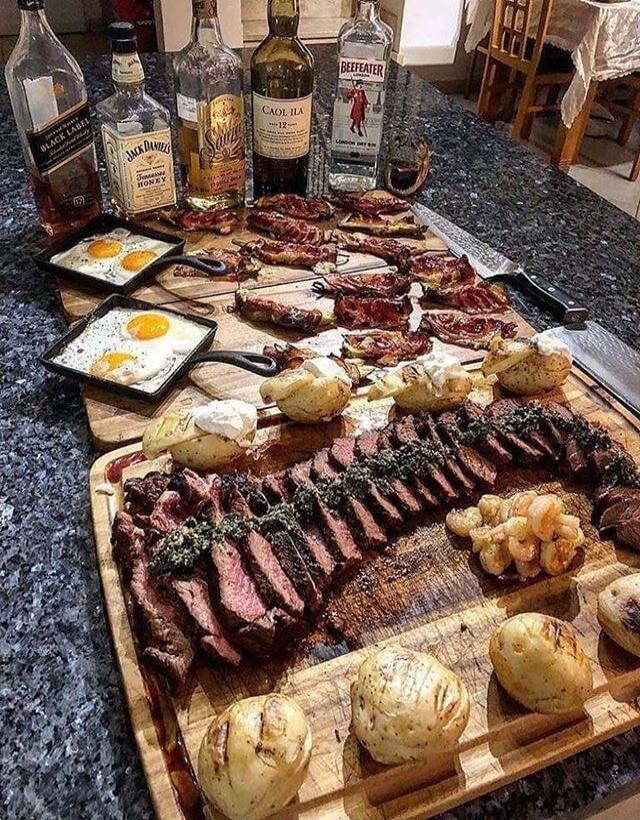 Bottle labels visible: Jack Daniel's, Beefeater, Caol Ila (Islay), Beefeater Gin. On the counter: a large wooden board with sliced steak, roasted potatoes, and some charred garlic or onions. Small pans with fried eggs over easy. The scene includes multiple bottles of liquor and a spread of meat (steak) and bread rolls on a granite countertop.