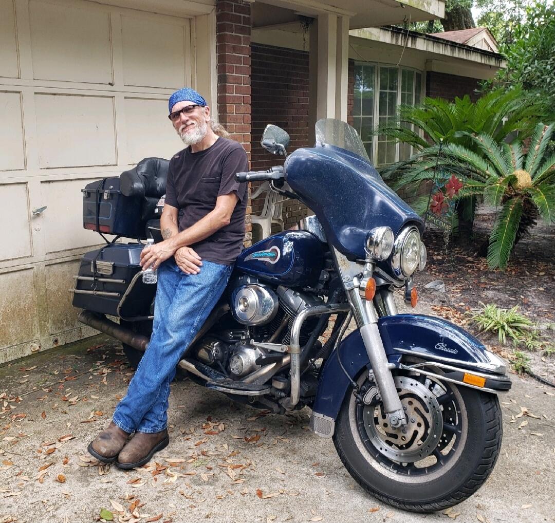 A man with a bandana and beard is leaning against a blue motorcycle. He is wearing a black t-shirt, jeans, and boots, holding a water bottle. A 'WELCOME' sign is visible in the background.