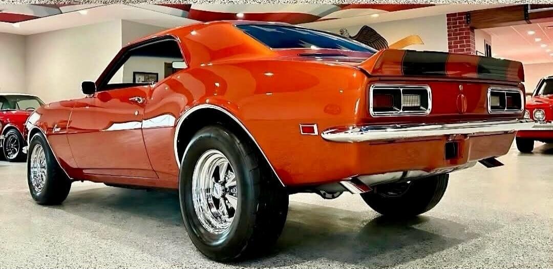 A classic orange muscle car, possibly a Chevrolet Camaro, is parked indoors. The car has a sleek design with chrome accents and racing stripes on the rear spoiler.