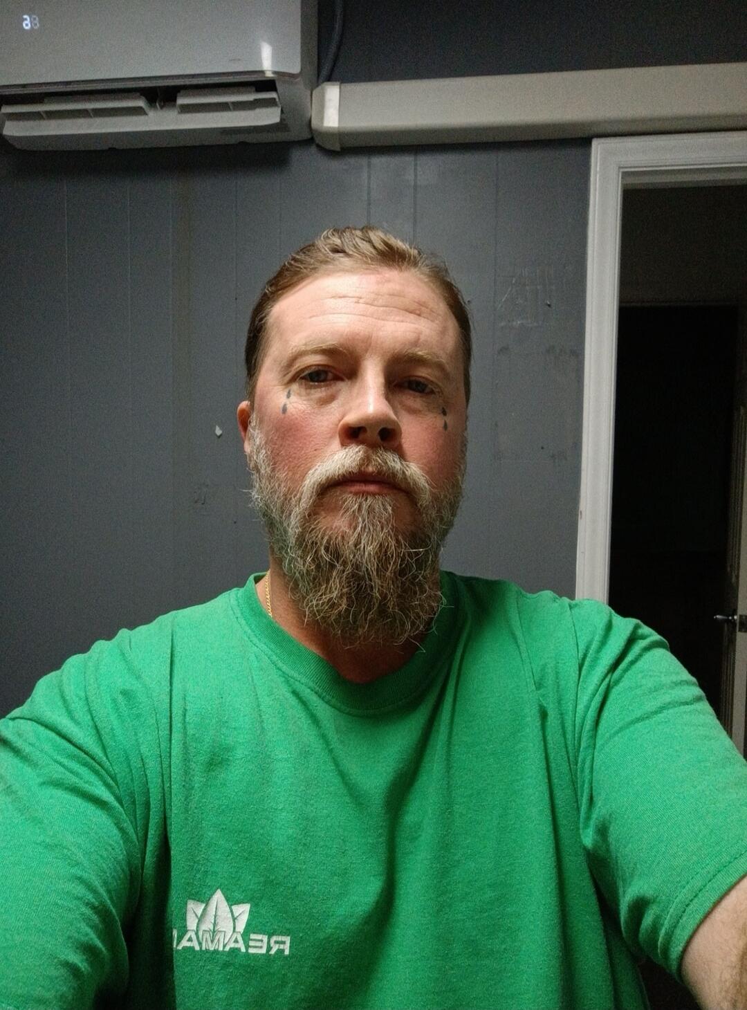 A man with a beard and teardrop tattoos under his eyes. He is wearing a green t-shirt with 'AMAZA' (reversed) text. An air conditioner unit in the background shows '38'.