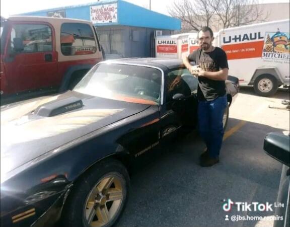 A man stands next to a black classic car, possibly a Pontiac Firebird Trans Am, with gold accents. In the background, there's a red Jeep, a blue building with 'DONALD R. DAVIS CLEANING & REPAIR' visible, and several U-Haul trucks and trailers. The car has 'T/A' on its hood scoop. The image also shows a TikTok Lite watermark with '@jbs.home.repairs'