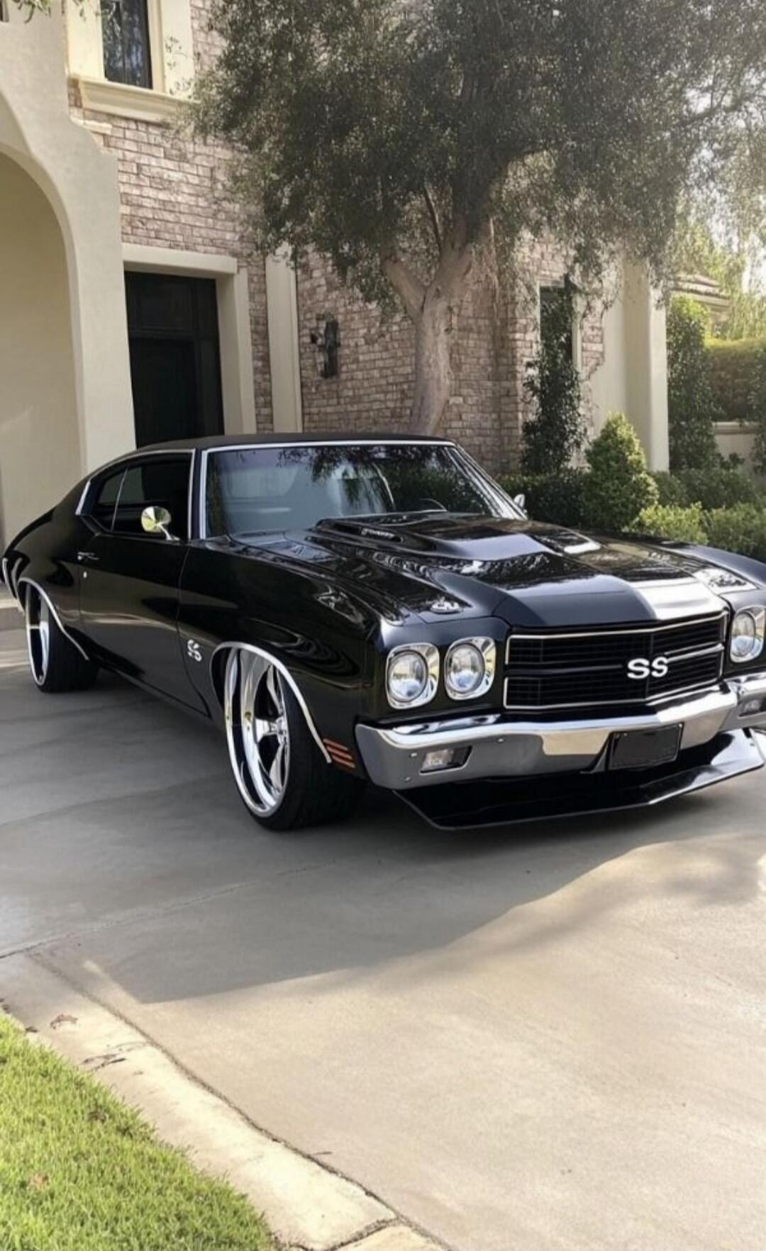 SS on front grille of a black muscle car parked in a suburban driveway. The car has chrome bumper, large white-lettered wheels, and a sleek black paint job. The house and trees are in the background.