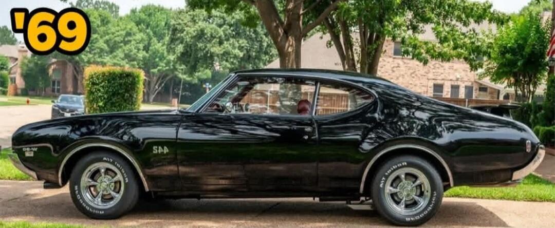 A black classic muscle car, model year '69, parked on a suburban street. The car has 'SAA' written on its side and 'BFGoodrich' tires.