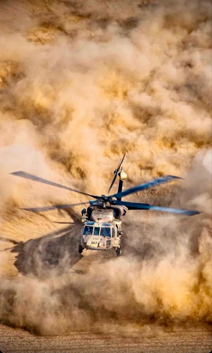 A military helicopter, possibly an HH-60 Pave Hawk with the number 567 visible, is flying low over a desert landscape, kicking up a large cloud of dust and sand.