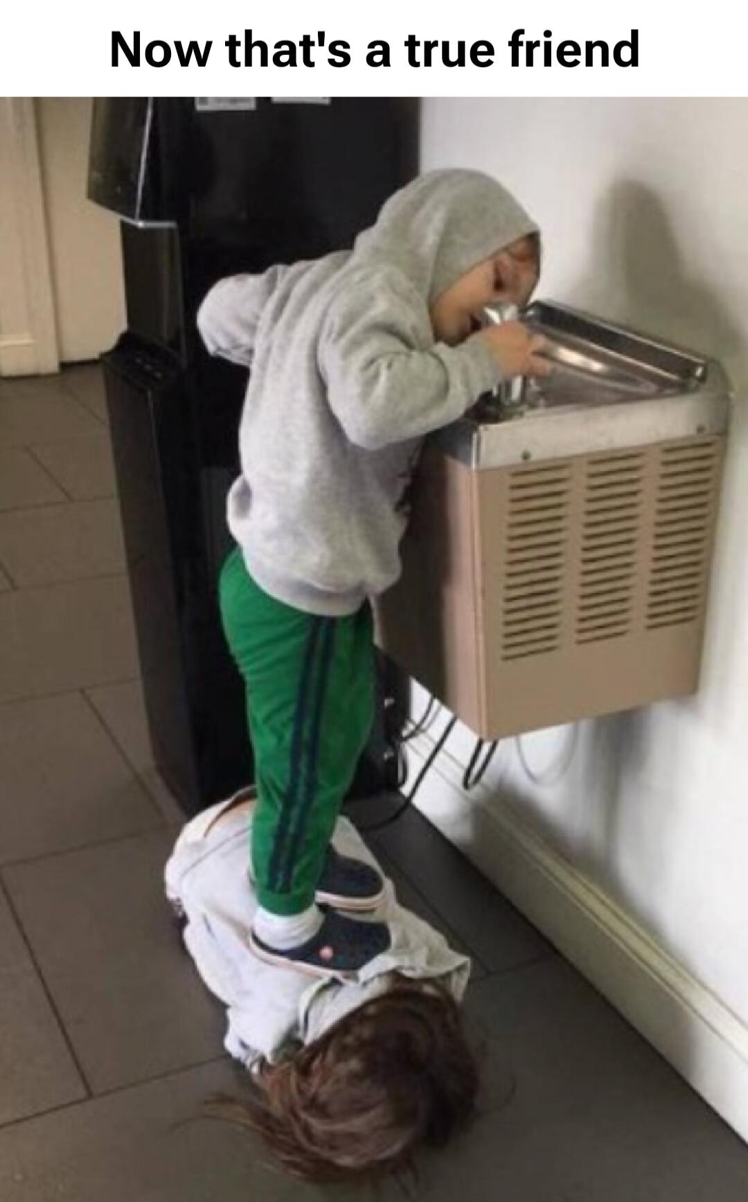 A child is standing on another child's back to reach a water fountain. The text on the image reads: 'Now that's a true friend'.