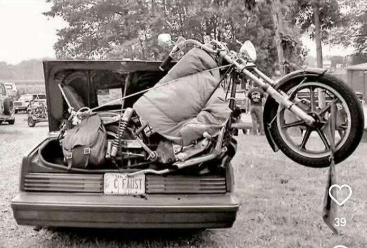 License plate: CAUST (hard to read). A vintage motorcycle is strapped inside the open trunk of a car in a black-and-white photo.
