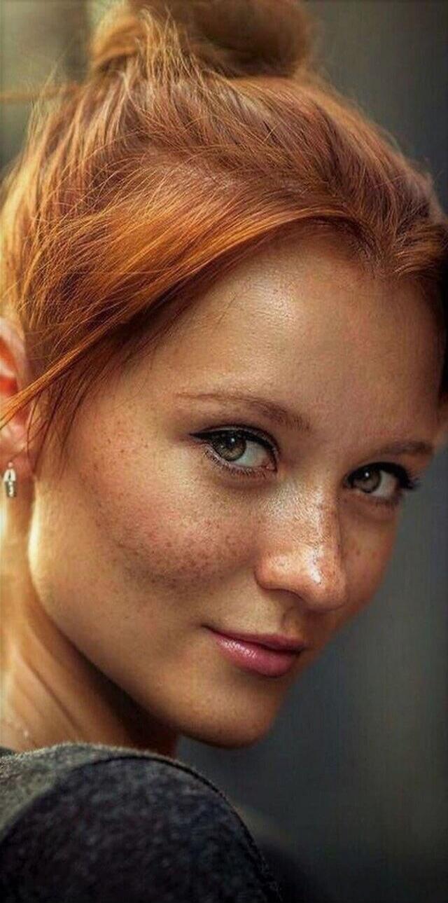 A close-up portrait of a young woman with red hair and freckles, looking over her shoulder with a soft smile.