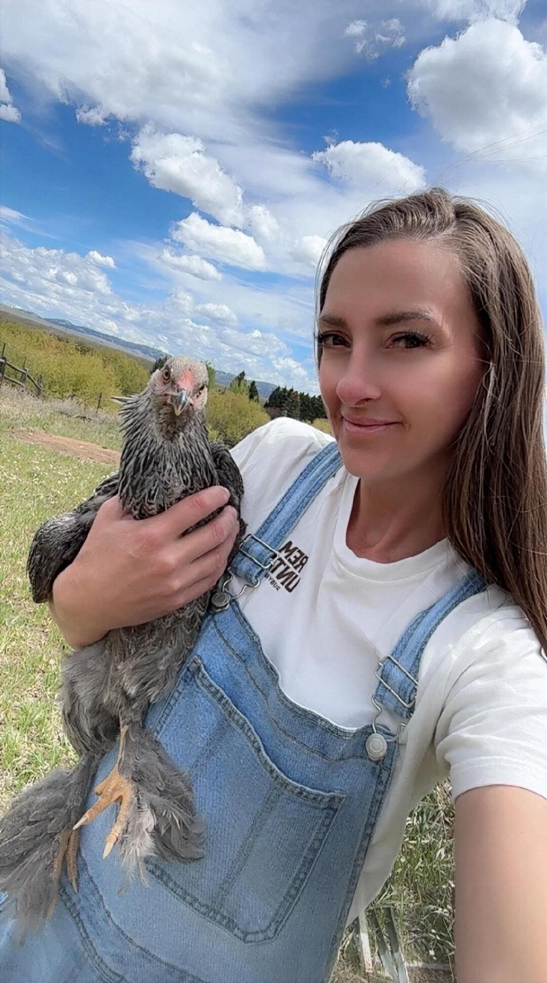 A woman in denim overalls is holding a chicken in a sunny outdoor setting.