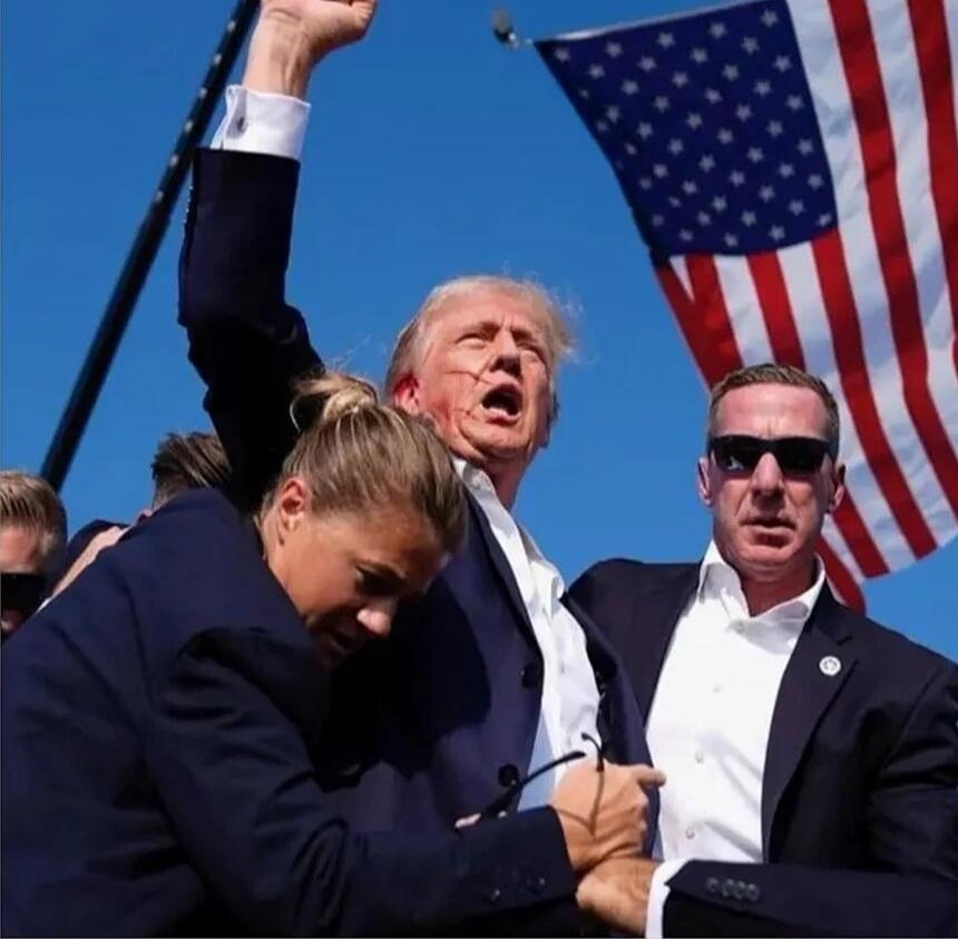 A political rally scene showing a man in a suit raising his fist, flanked by security and supporters, with an American flag in the background.