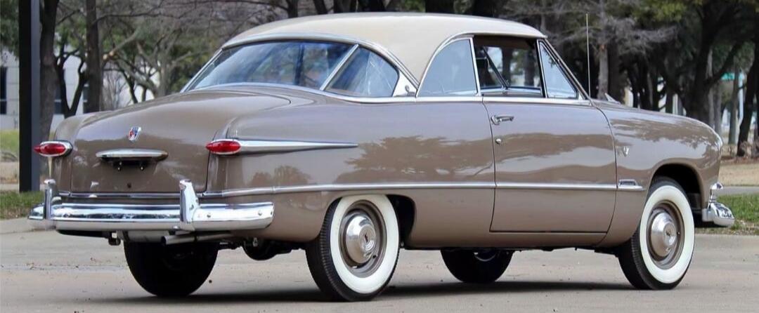 A vintage two-door coupe car, light brown with a cream-colored roof, parked outdoors. It features classic chrome details and whitewall tires.