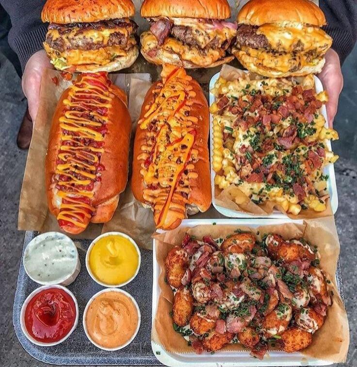 A tray of assorted fast-food items: three large burgers with buns, two large hot dogs topped with sauces, a tray of loaded fries with toppings, and several dipping sauces (ranch, yellow mustard, red ketchup, and an orange sauce).
