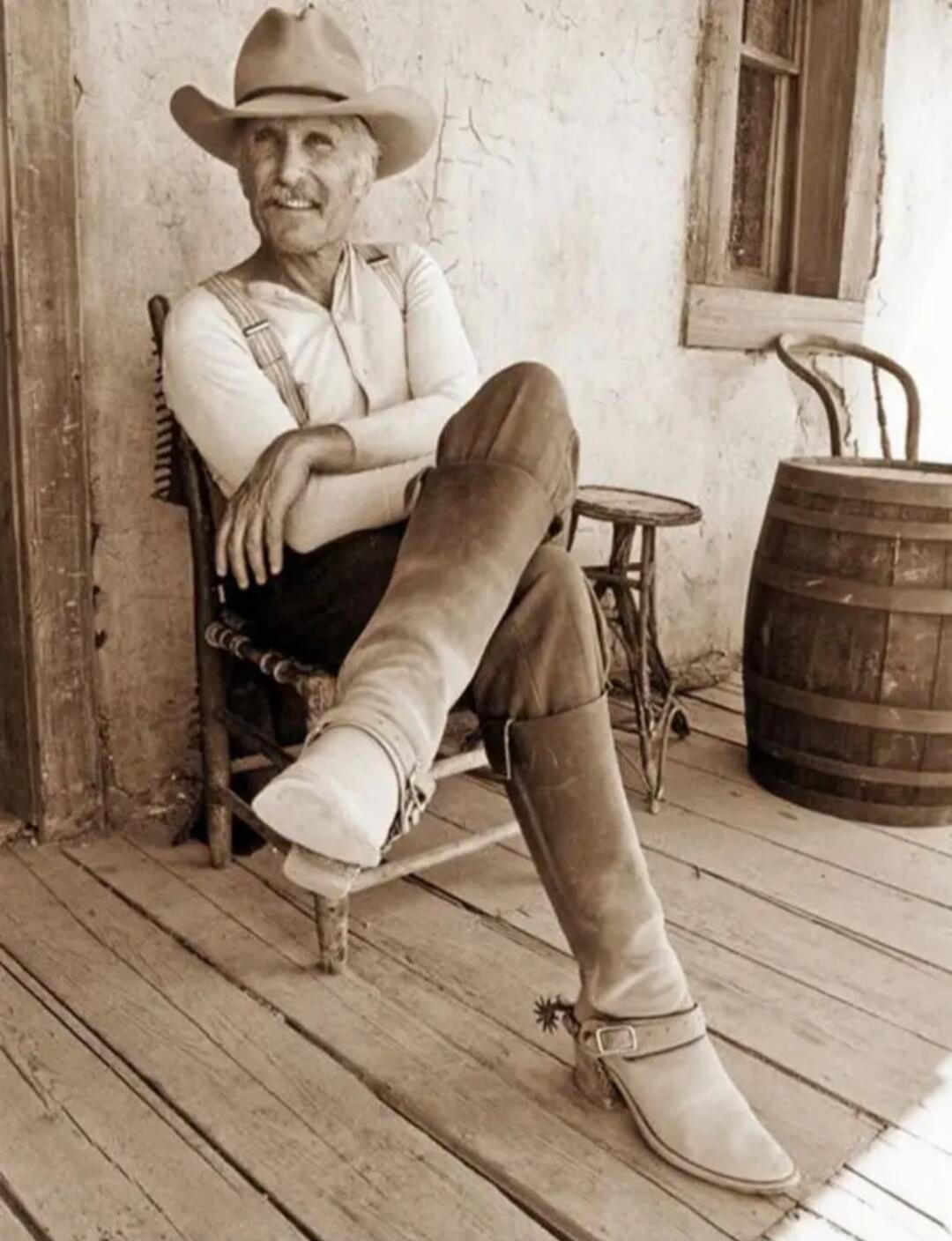 An elderly man wearing a cowboy hat sits on a wooden porch, his legs crossed, wearing boots and a light shirt with suspenders. A wooden barrel and a chair are nearby, and he smiles at the camera.