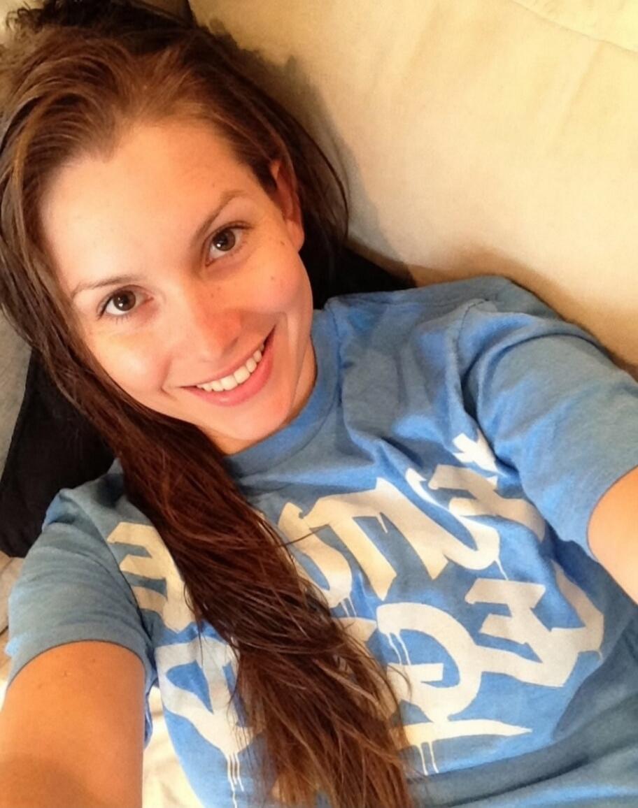A young woman is taking a selfie, smiling at the camera. She has long brown hair and is wearing a light blue t-shirt with a white graphic design.