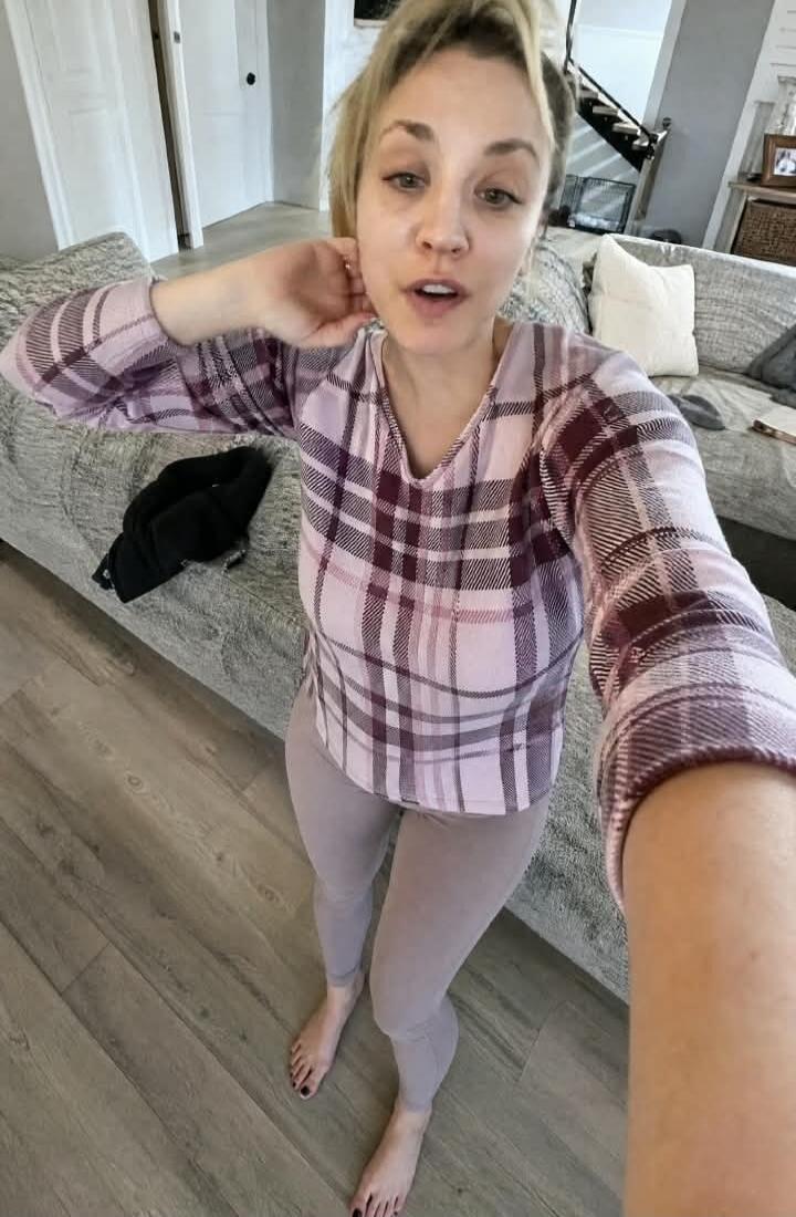 A woman, possibly Kaley Cuoco, takes a selfie indoors. She is wearing a plaid long-sleeve shirt and light-colored leggings, standing barefoot on a wooden floor. Her hair is tied up, and she appears to be without makeup. A couch is visible in the background.
