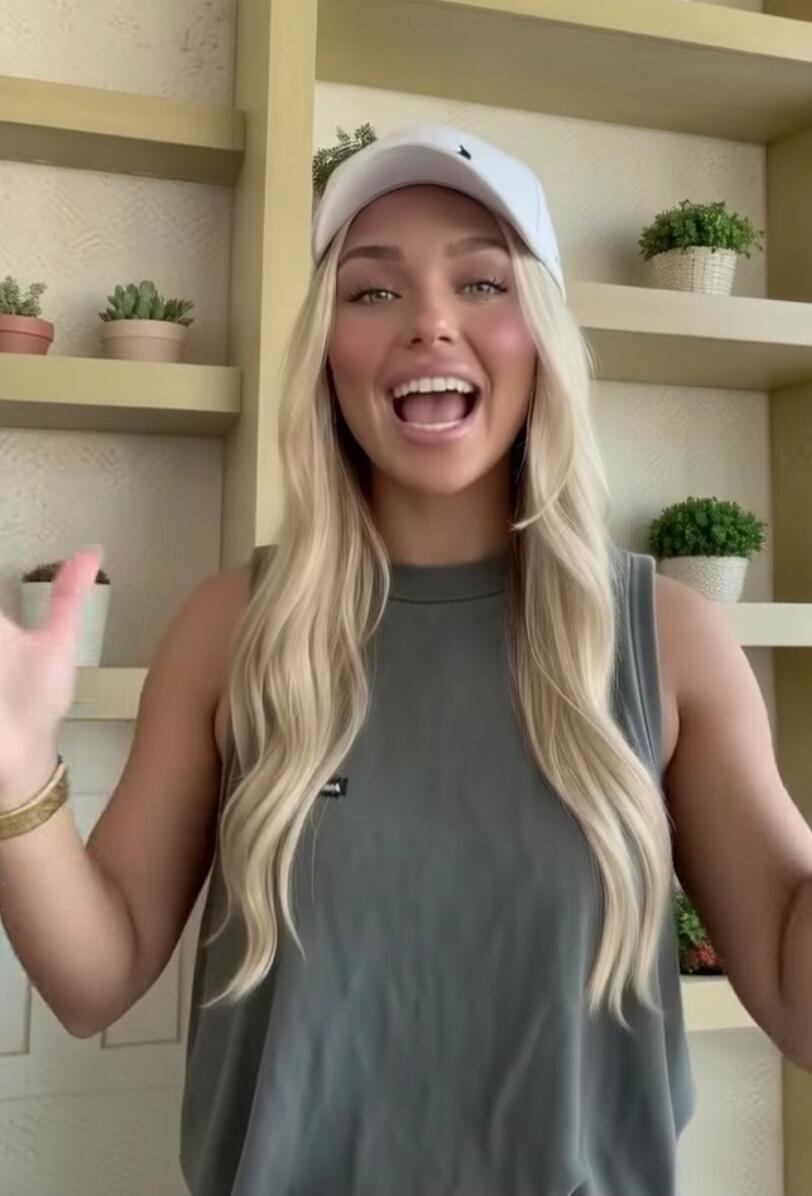 A young blonde woman wearing a white baseball cap and a grey tank top smiles broadly at the camera, gesturing with her hands. She is standing in front of shelves with potted plants.