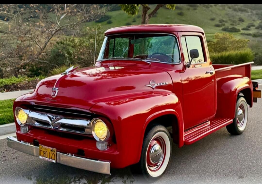A bright red vintage Ford pickup truck is parked on an asphalt road with a green, hilly background. The truck has chrome accents, white wall tires, and a California license plate C80 123.