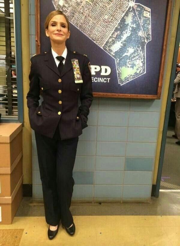 A woman in a dark blue police uniform, complete with a tie and medals, stands in front of a map with 'PD ECINCT' visible. She is smiling slightly with her hands in her pockets.