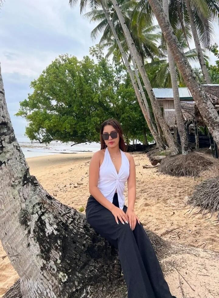 A young woman is sitting on a fallen palm tree trunk on a sandy beach, wearing a white halter top and black pants. Palm trees and the ocean are visible in the background.