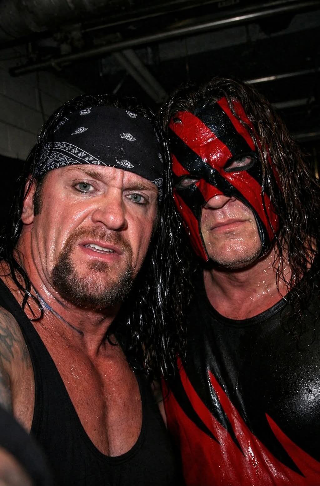 Two muscular men posing for a photo; one wears a black bandana and tank top, the other wears a black and red mask with matching attire, both appearing sweaty.