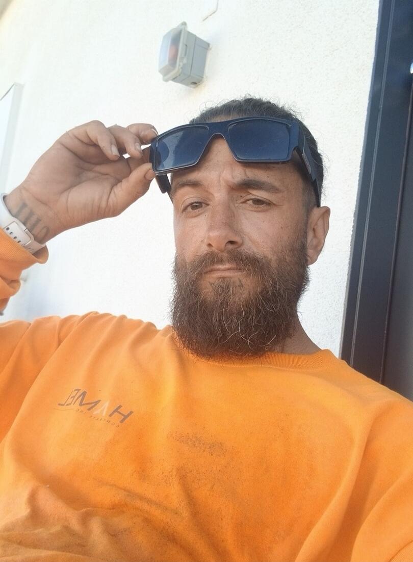 A man with a beard is wearing an orange long-sleeve shirt and holding sunglasses on his forehead. The shirt has mirrored text that appears to be 'HWE CONCRETE INC'. There is a tattoo on his wrist that looks like Roman numerals 'IIII'.