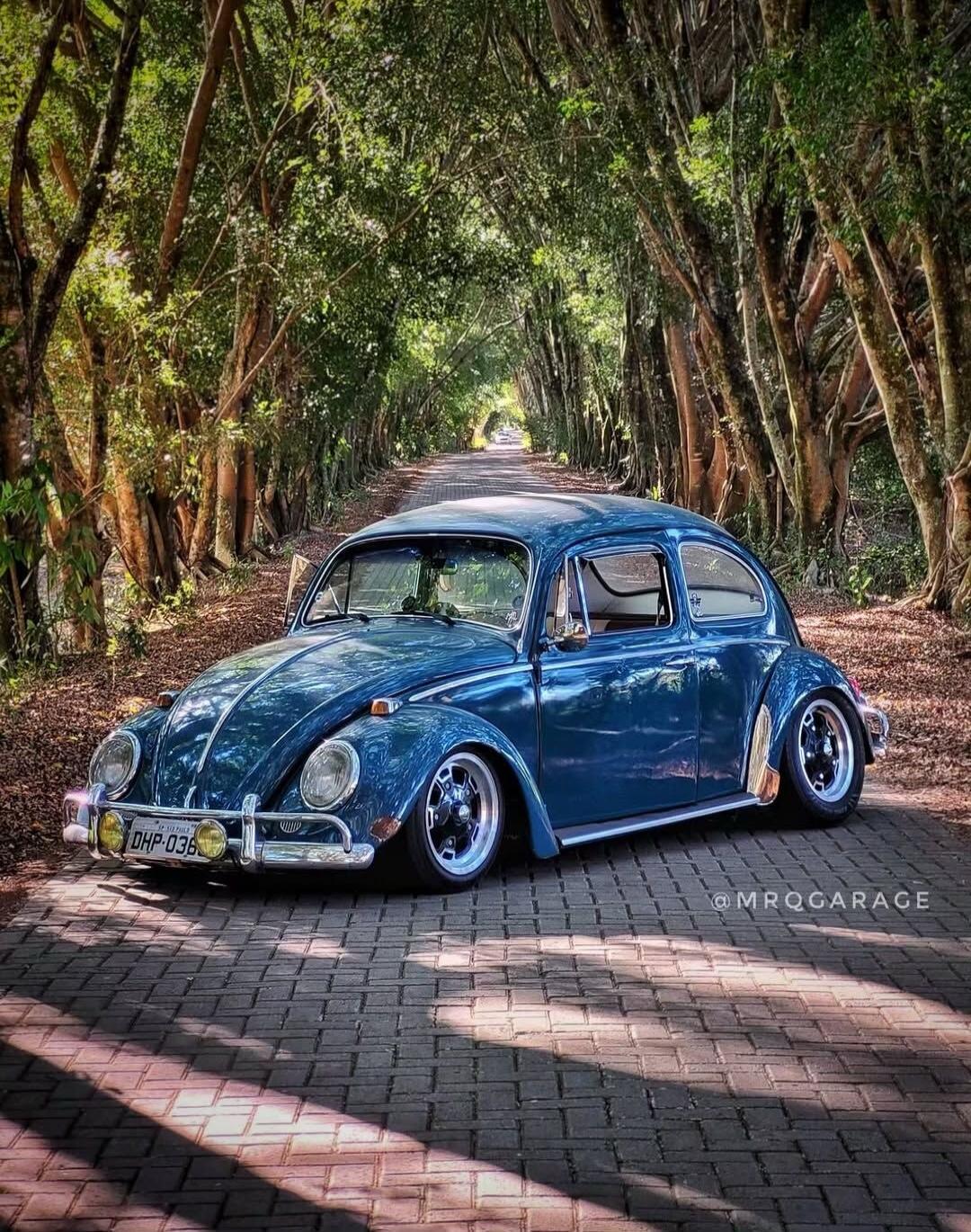 The image shows a blue Volkswagen Beetle parked on a brick road, surrounded by a tunnel of trees. The car is classic and appears to be modified with lowered suspension and custom wheels. The lighting suggests it's daytime, with sunlight filtering through the leaves, creating dappled shadows on the road. There is a license plate visible on the front