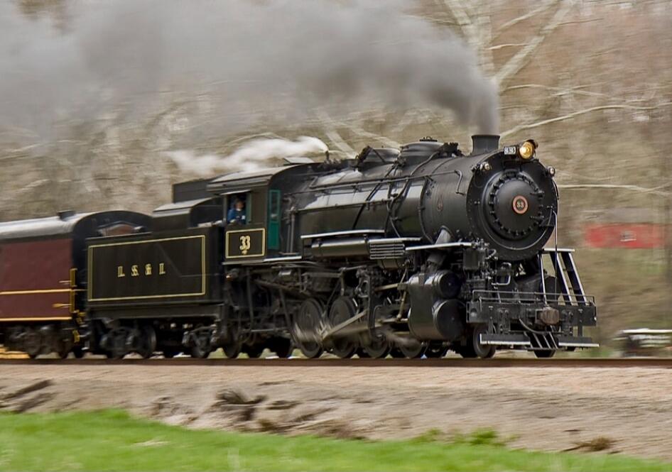 A black steam locomotive pulling a coal tender, emitting smoke as it moves along the track.