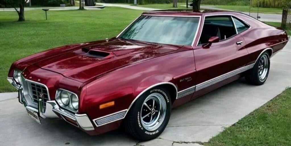 A classic maroon Ford Torino muscle car is parked on a paved driveway with green grass in the background.