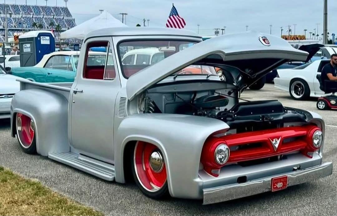 A custom silver pickup truck with red accents, including the grille and wheels, parked at an outdoor event. The hood is open, revealing the engine. An American flag is visible in the background, along with stadium seating and other vehicles. The license plate reads 'GREASY NUTS 53'.