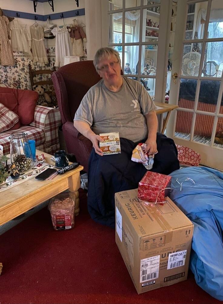 A person wearing a t-shirt with 'the YMCA' logo is sitting in an armchair, holding two small boxes marked '21'. A large cardboard box from 'KEURIG K-SELECT' is on the floor, with a red wrapped gift on top. The box also has 'FRAGILE' and 'UPC 0877' visible. The room is a cozy living space with various decorations and furniture.