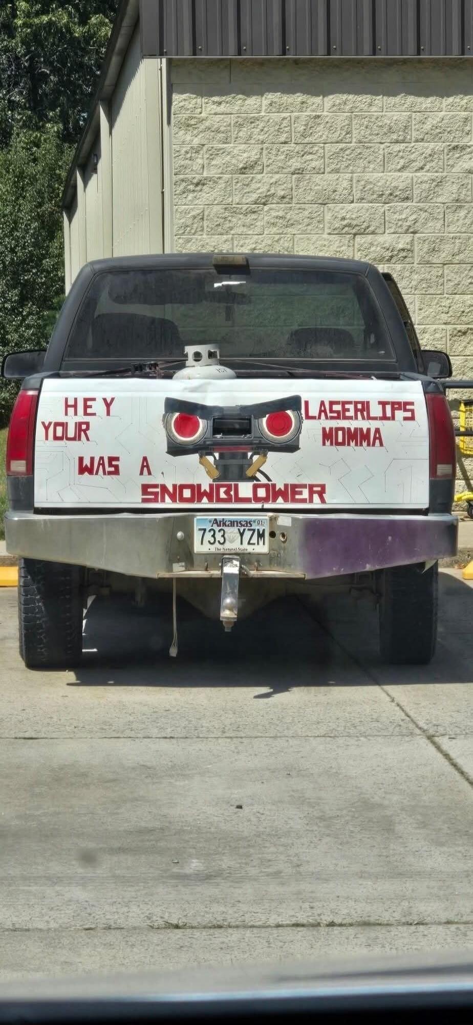 HEY YOUR LASERLIPS MOMMA WAS A SNOWBLOWER. This text is painted on the tailgate of a pickup truck, which also features a custom face-like design with red eyes and a propane tank in the truck bed. The license plate reads 'Arkansas 01 733 YZM, The Natural State'.