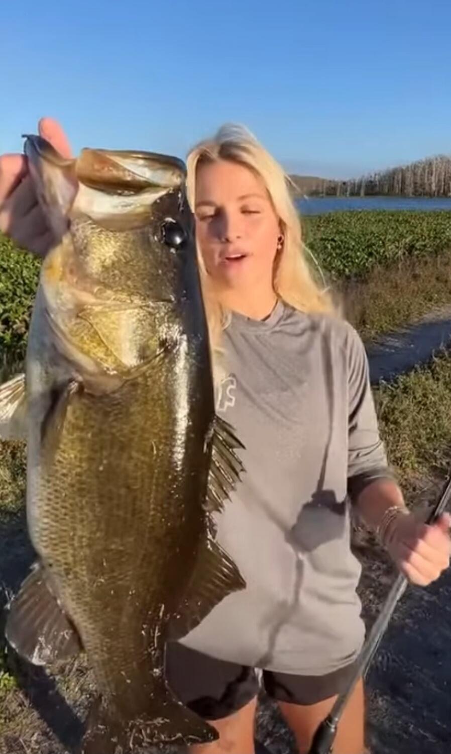 A woman holding a large fish outdoors near a body of water.