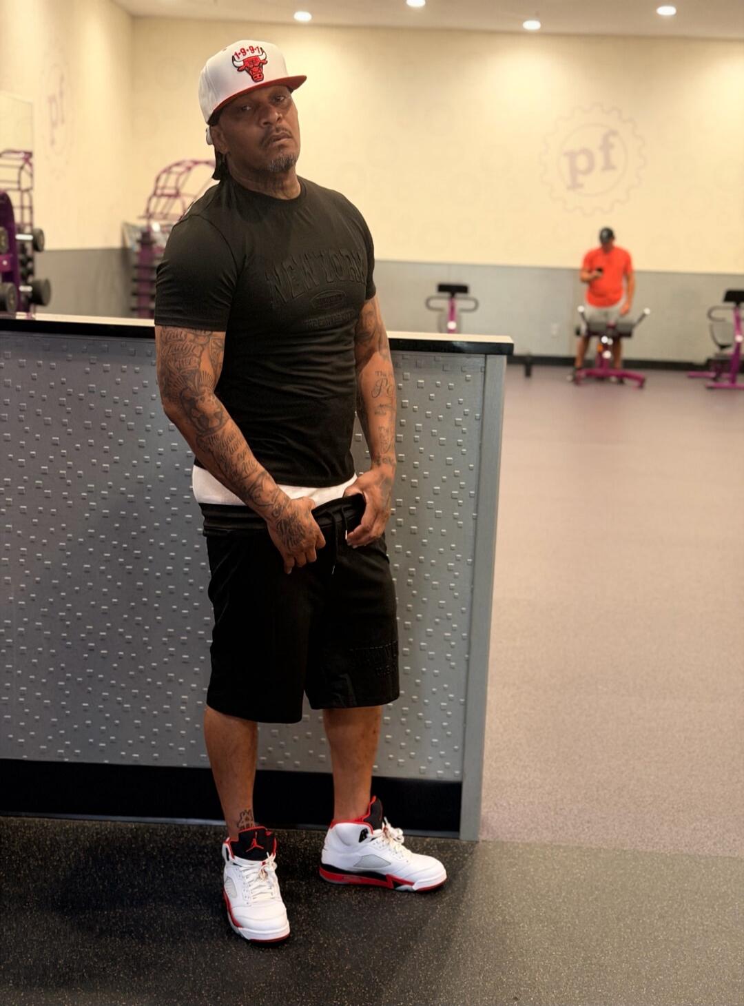 A man in a gym wearing a black t-shirt, black shorts, a white undershirt, a white and red Chicago Bulls cap, and white and red sneakers. He has tattoos on his arms. Another person is visible in the background. The gym has a 'pf' logo on the wall.