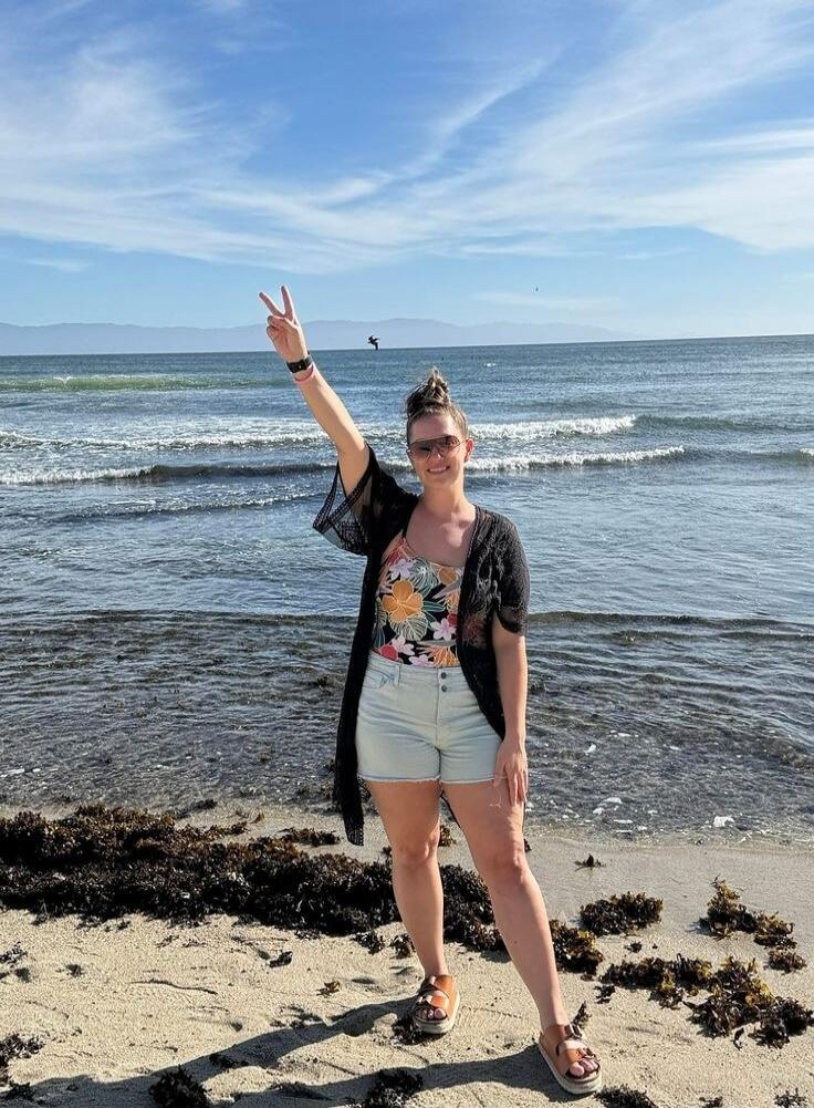 A woman stands on a sandy beach, smiling and making a peace sign with her right hand. She is wearing a floral swimsuit, light shorts, a black sheer cover-up, sunglasses, and sandals. The ocean with gentle waves is behind her, under a clear blue sky with some clouds. Seaweed is visible on the wet sand.