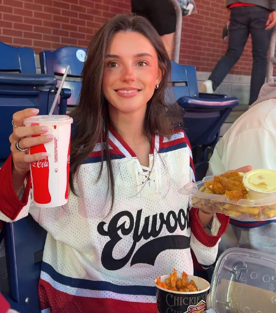A young woman wearing a white, red, and blue jersey with 'Elwood' written on it, holding a Coca-Cola cup and a tray of chicken tenders and fries. Another cup with fries is visible. The background shows stadium seats.