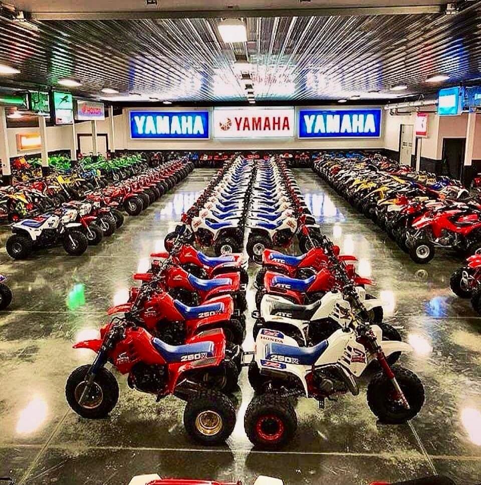 YAMAHA YAMAHA YAMAHA showroom filled with rows of small motorcycles/ATVs. Bright banners with 'YAMAHA' along the back wall. The floor is polished and reflects the overhead lighting. Numerous red, blue, and white mini motorcycles/ATVs are neatly lined in symmetrical rows.