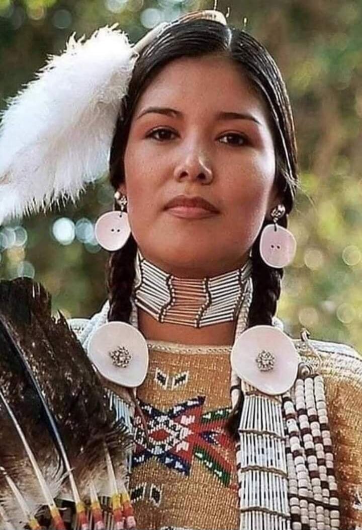 A young woman in traditional attire with beaded jewelry and large circular white ornaments, braided hair, and a feather headdress piece.