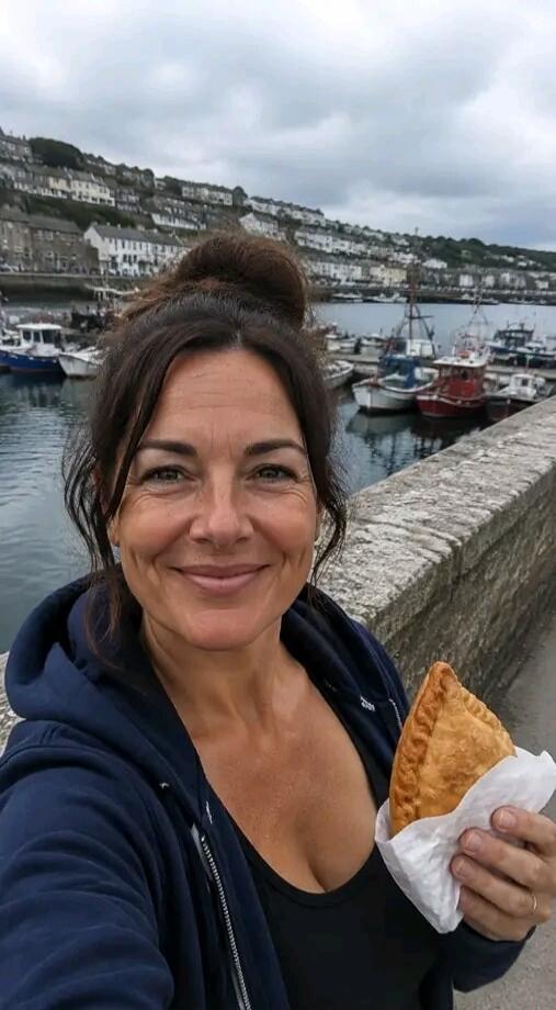 A woman smiles at the camera, holding a pasty. In the background, there is a harbor with boats and houses on a hillside.