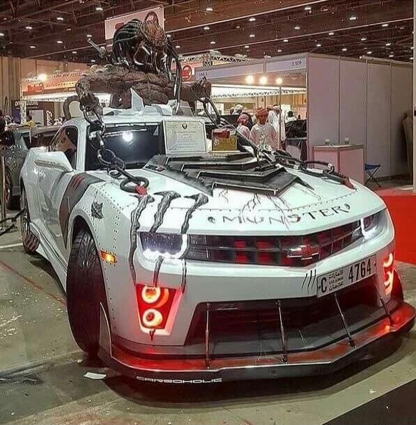 A heavily customized white Chevrolet Camaro with a monster theme, featuring a large creature sculpture on the roof, chains, rivets, and red accents. The word 'MONSTER' is visible on the hood, and 'CARBOHOLIC' on the front spoiler. The car is displayed at an indoor exhibition.