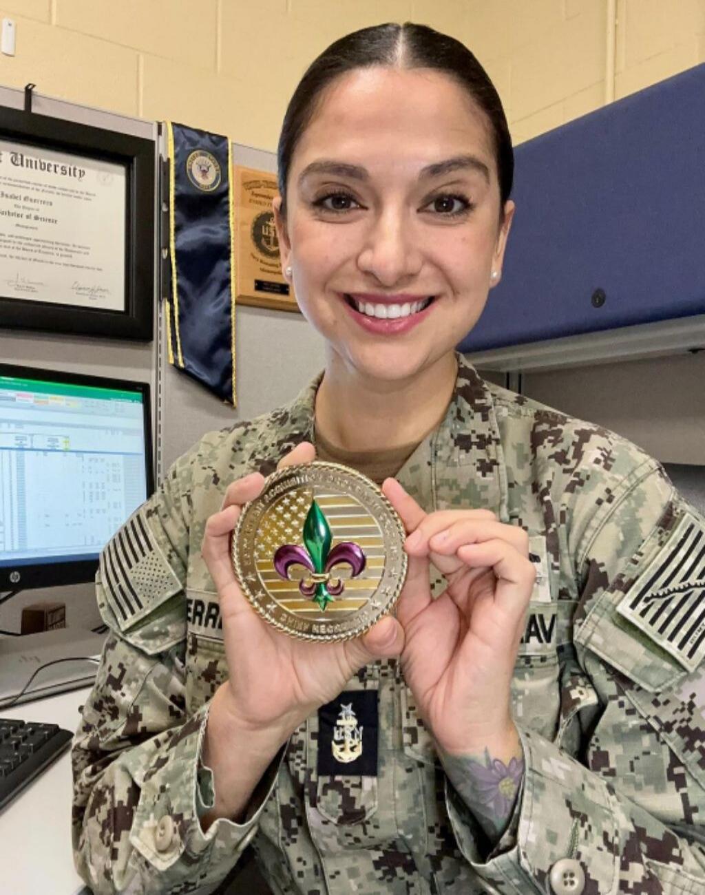 A smiling female soldier in camouflage uniform proudly holding a military medal featuring a green fleur-de-lis design.