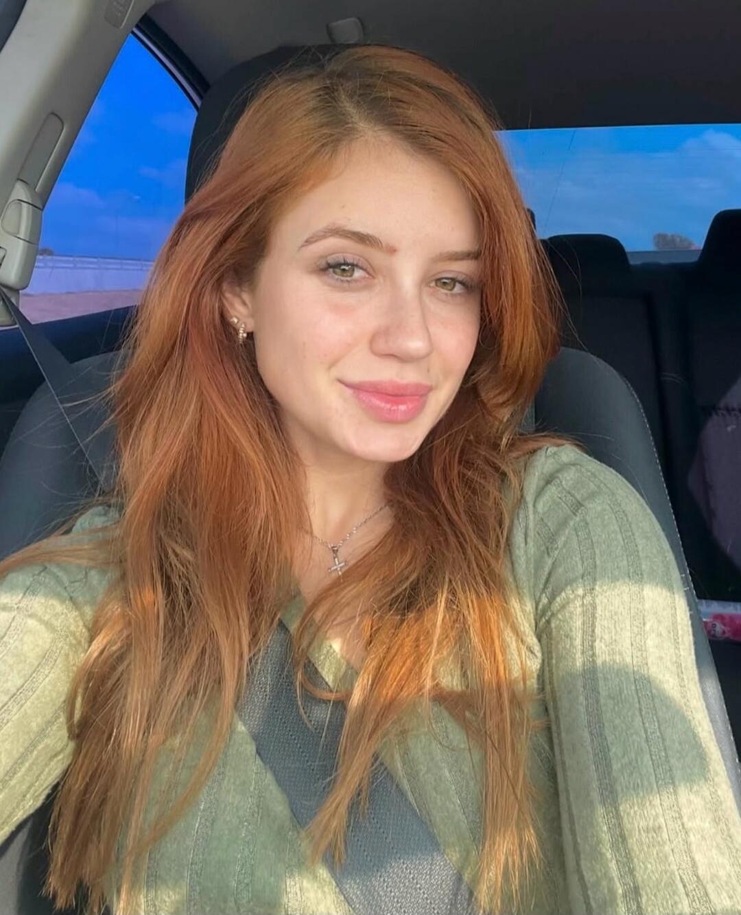 A young woman with long red hair and a green top is smiling at the camera from inside a car, wearing a seatbelt.