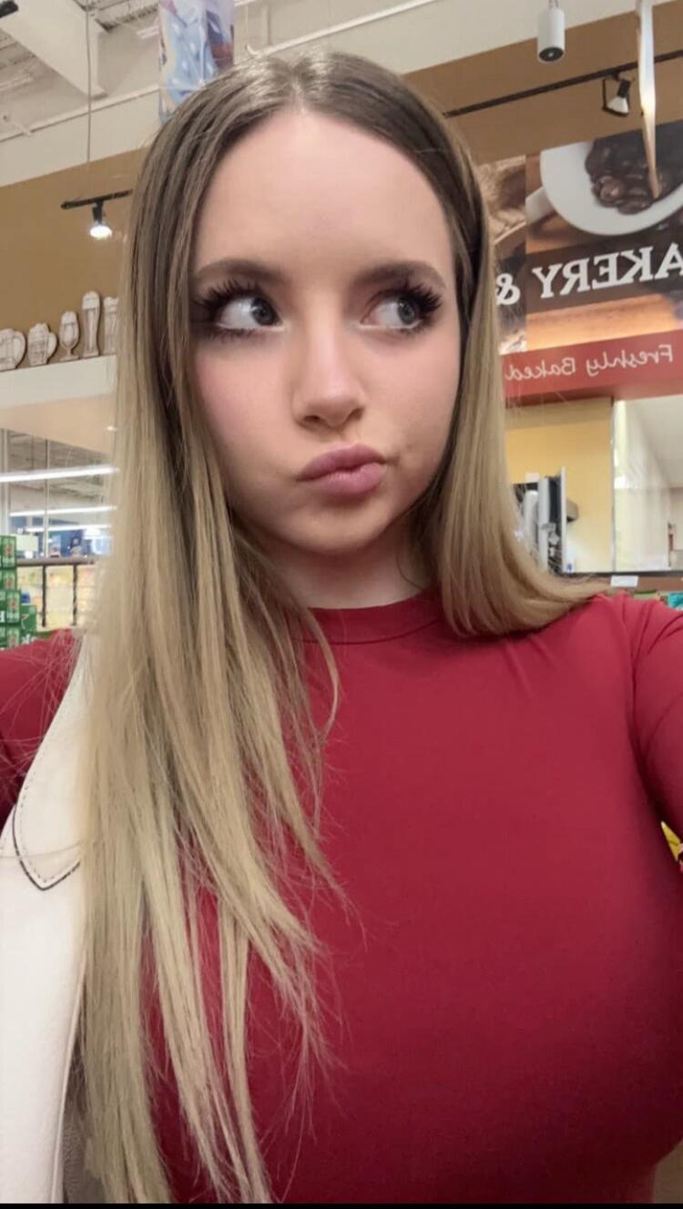 A young woman with long blonde hair and a red top, making a pouty face and looking to the side in what appears to be a supermarket. Text visible in the background includes 'BAKERY' (reversed).