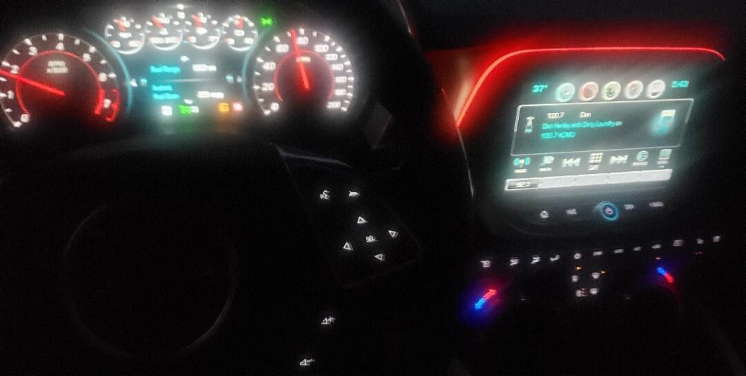 Dashboard shows RPM, fuel range, and speed up to 200. The infotainment screen displays 37 degrees, current time 5:43, and radio station 100.7 playing 'Dan Harley with Dirty Laundry on 100.7 KMD'. Steering wheel buttons include 'SEL'.