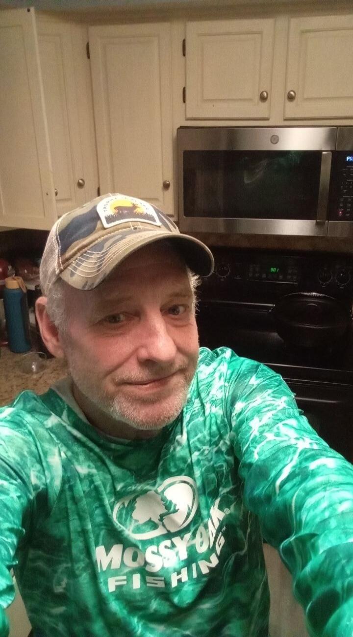 A man wearing a green long-sleeve shirt with 'MOSSY OAK FISHING' logo and a camouflage baseball cap, smiling at the camera in a kitchen setting.