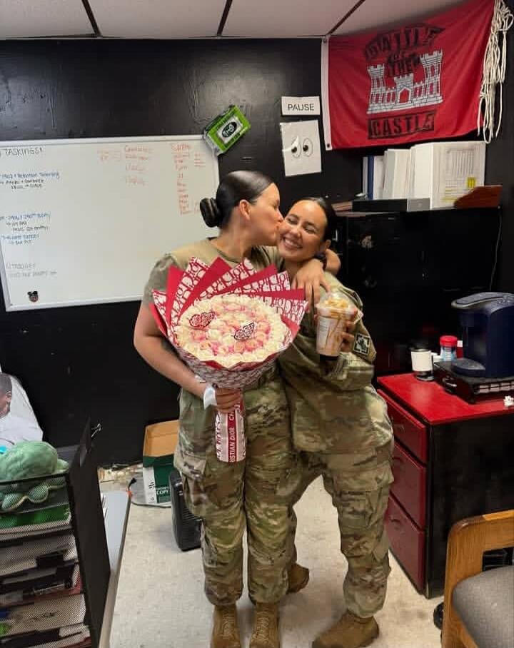 Two women in military uniforms are hugging. One woman is kissing the other on the cheek. The woman being kissed is holding a drink, and the other is holding a large bouquet, possibly a candy bouquet, wrapped in paper with 'CHRISTIAN DIOR' branding. Both are smiling. In the background, there's a whiteboard with 'TASKINGS', a red flag with 'BATTLE OF
