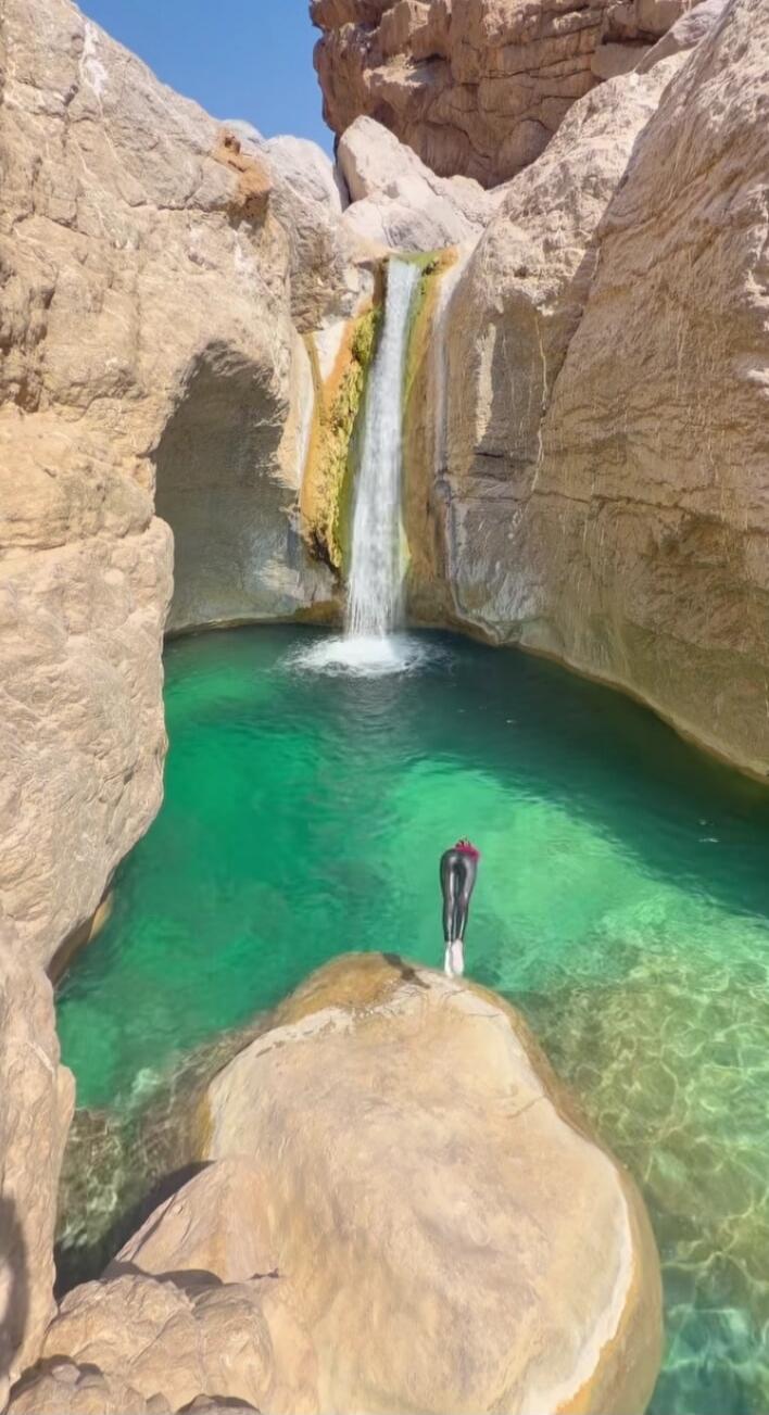 A person in a wetsuit stands on a rock overlooking a beautiful waterfall cascading into a clear turquoise pool in a rocky canyon.
