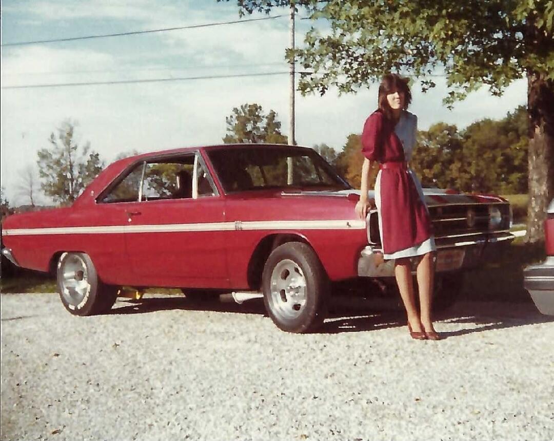 A vintage photograph showing a woman standing next to a classic red two-door car, possibly a Dodge Dart, on a gravel surface. The woman is wearing a two-tone dress and leaning against the front fender of the car. Trees and a clear sky are visible in the background.
