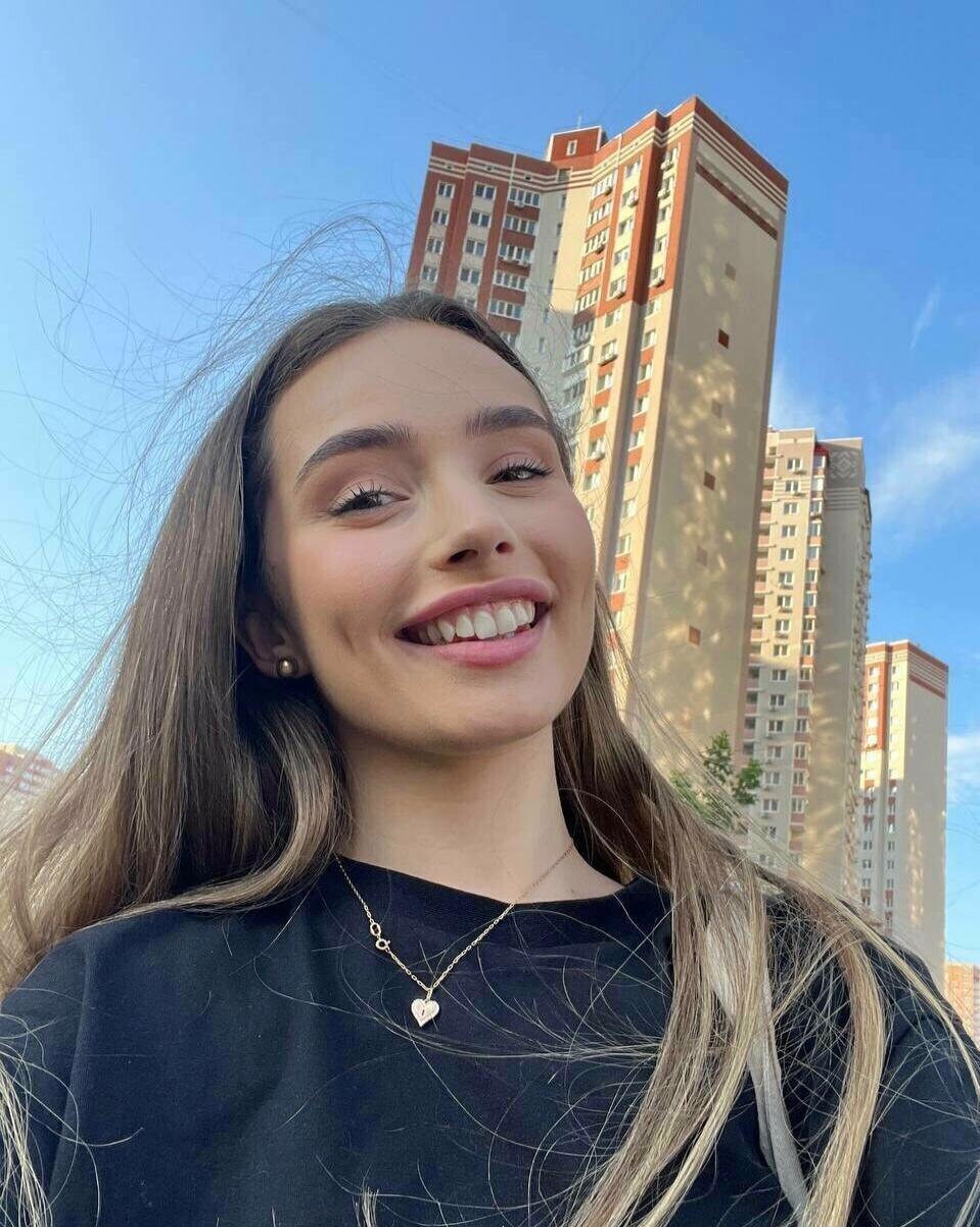 A young woman with long brown hair smiles broadly at the camera, taking a selfie. She is wearing a black top and a delicate gold necklace with a heart-shaped pendant. In the background, tall modern residential buildings with red and beige facades stand against a clear blue sky.