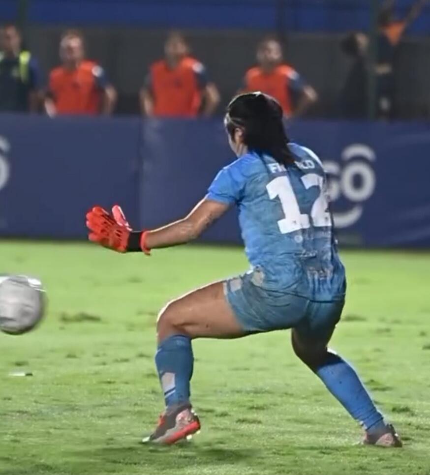A female soccer goalkeeper, wearing a light blue jersey with number 12 and 'FRANCO' on the back, and red gloves, is in a crouched position on a green field, ready to play the ball. A soccer ball is visible near her.