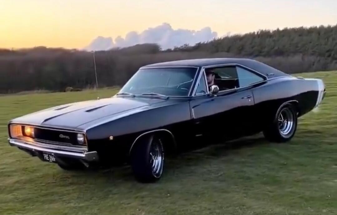 A black classic American muscle car parked on a grassy field. The car has a sleek body, wide tires, and a distinctive long hood with racing stripes.